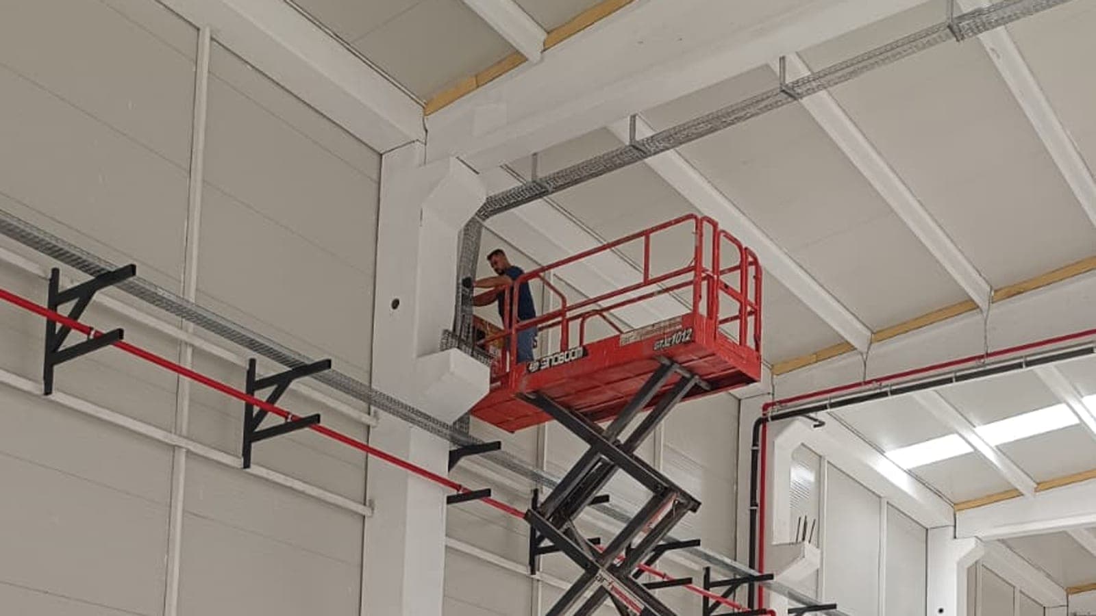 Kapalı üretim binasında akülü makaslı platformla çatı altı sprinkler hattı montajı yapan operatör